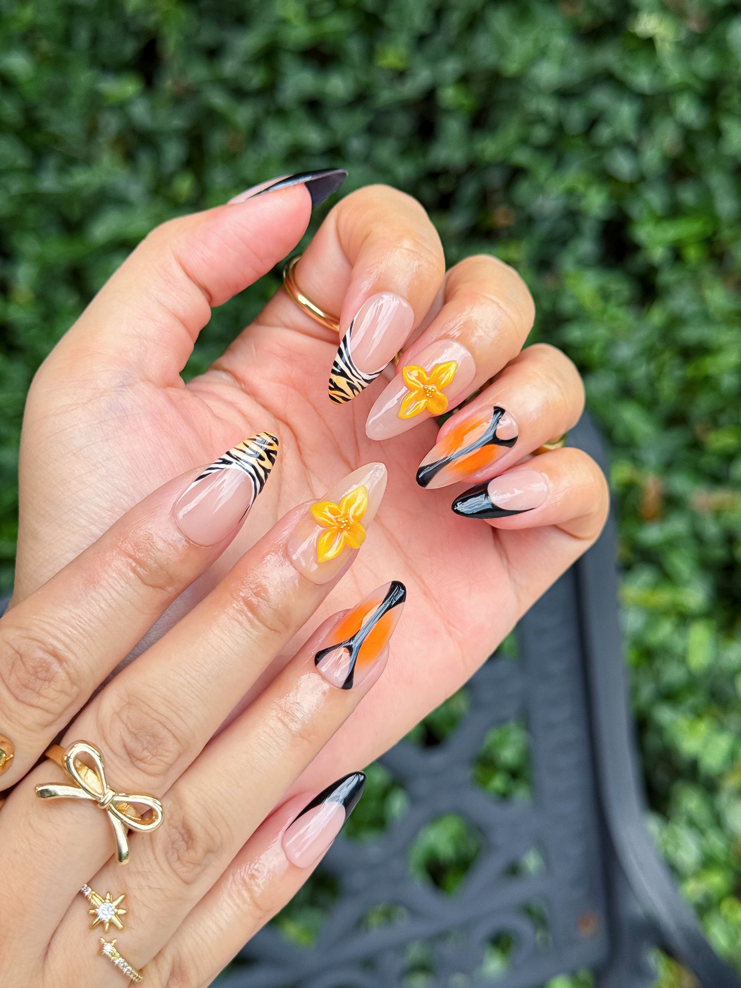 Deux mains avec manucure Tigre Fleurie, montrant des ongles en amande décorés de fleurs jaunes en 3D, motifs tigre et pointes noires, avec bijoux dorés en extérieur