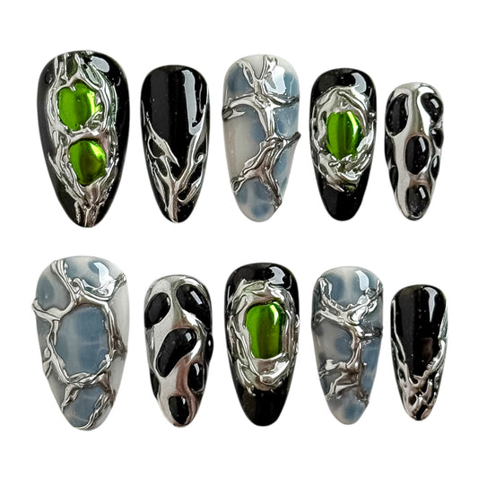Luxury press-on nails featuring biomechanical silver chrome textures, glowing green gemstone details and glossy black accents.