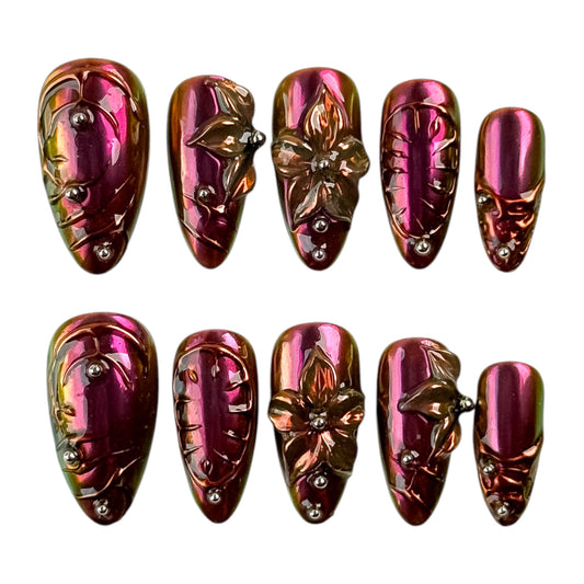 Manucure Amarante portée sur la main, ornée de fleurs en relief et de reflets métalliques aux nuances prune et bronze.