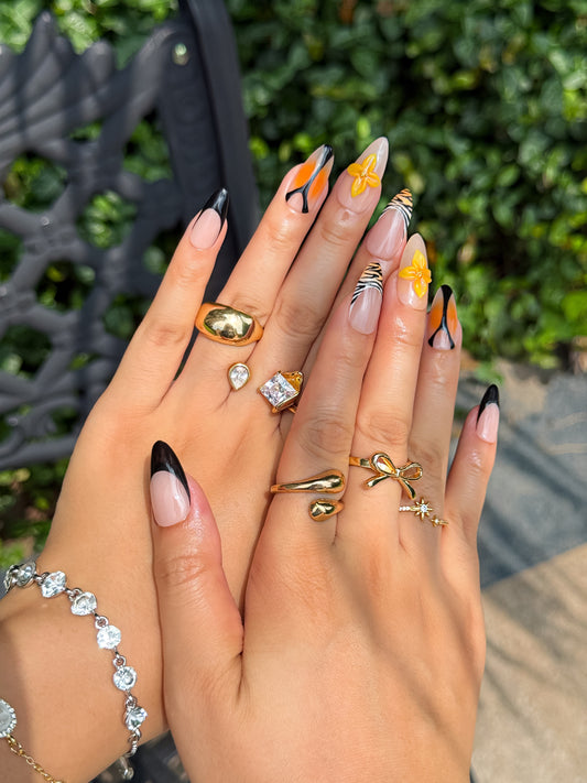 Main portant les ongles Tigre Fleurie, avec motifs tigre, fleurs 3D et détails noirs et orange, en extérieur avec fond de verdure